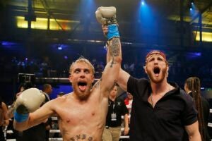 Jake Paul and Logan Paul, two light skin men, pose in a boxing ring. Logan holds his brother Jake's arm up - Jake is wearing boxing gloves and is shirtless, while Logan has a black shirt on. Jake opens his mouth, tongue out, while Logan also has his mouth open and wears a backward baseball cap.