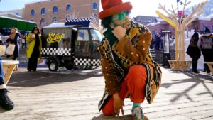 Boots Riley, a Black man, strikes a pose in colorful pants, coat, and tall hat, wearing sunglasses as he kneels on the boardwalk. A few people are behind him, including a small truck that reads 