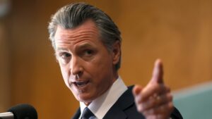 Gavin Newsom, a man with light-toned skin, holds up a finger, gesturing off camera emphatically, in front of a wall with wood paneling